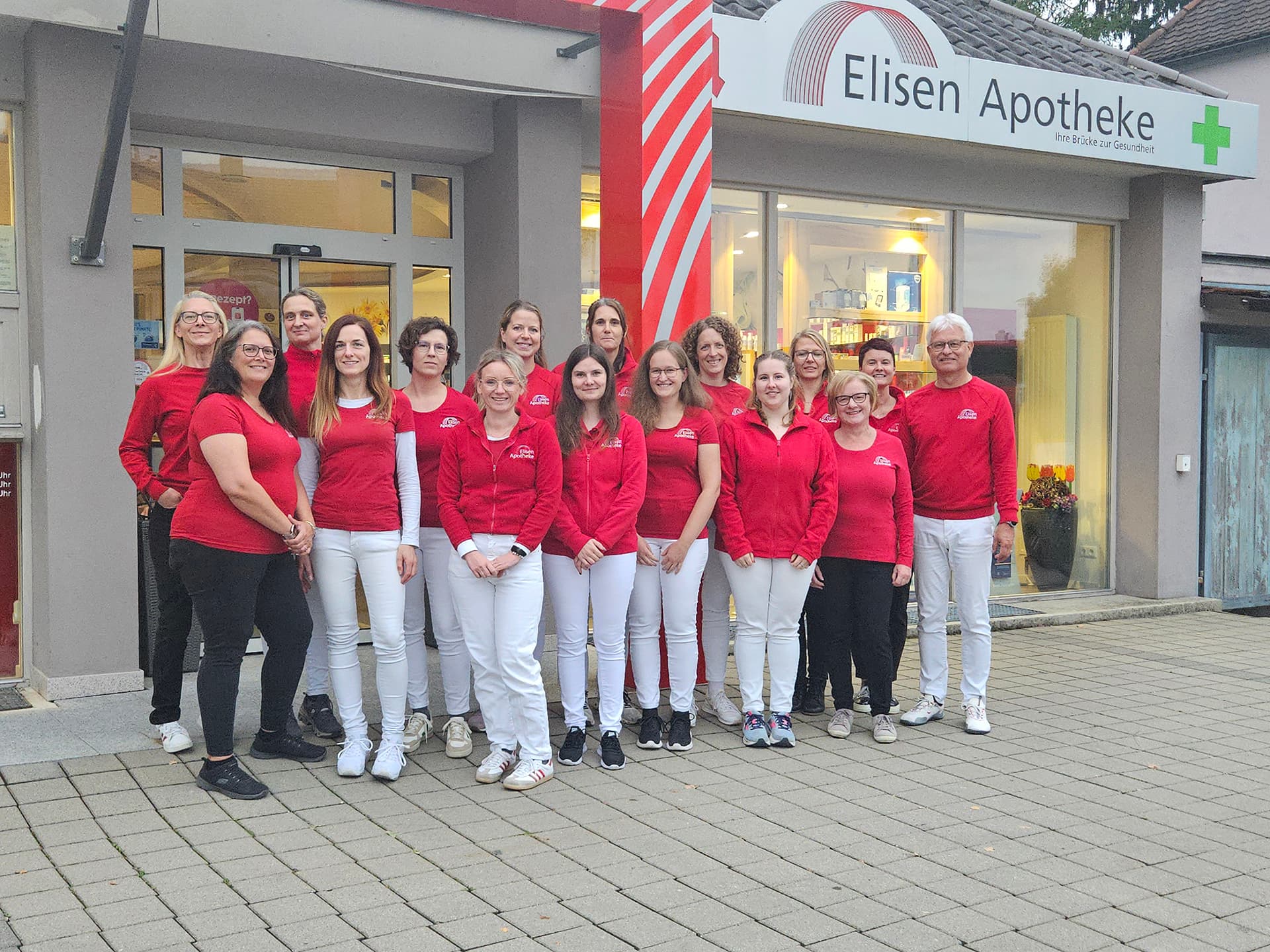 Mitarbeiterteam der Elisen Apotheke steht in roten Shirts vor dem Eingangsbereich.