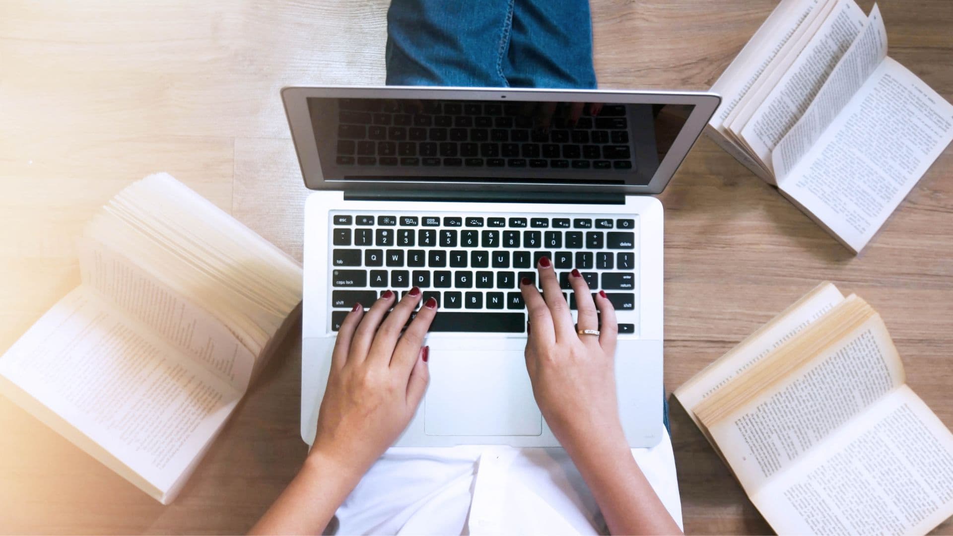 Hände tippen auf einer Laptop-Tastatur, umgeben von aufgeschlagenen Büchern auf einem Holztisch.